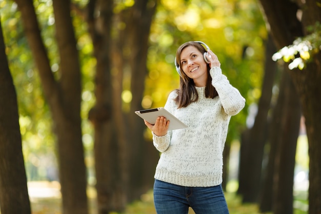 The beautiful happy cheerful brown-haired woman in white sweater with a tablet listening music in the white headphones in fall park on a warm day. Autumn in the city.