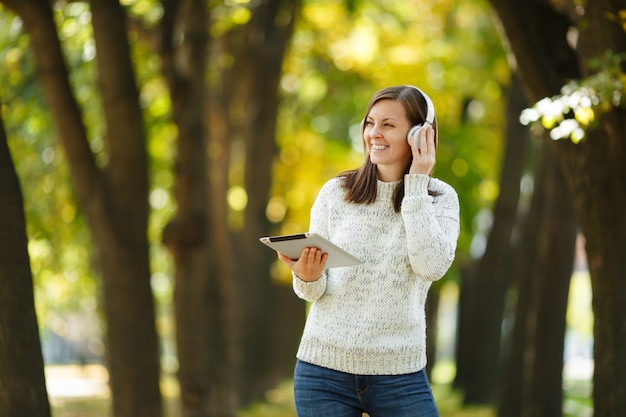 The beautiful happy cheerful brown-haired woman in white sweater with a tablet listening music in the white headphones in fall park on a warm day. Autumn in the city.