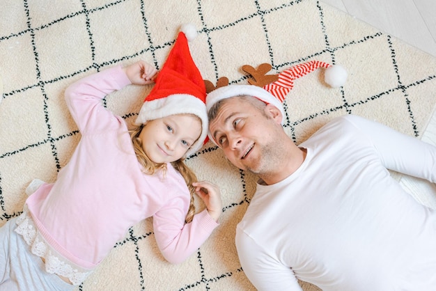 Beautiful happy family father and daughter in santa claus hats lie on rug floor