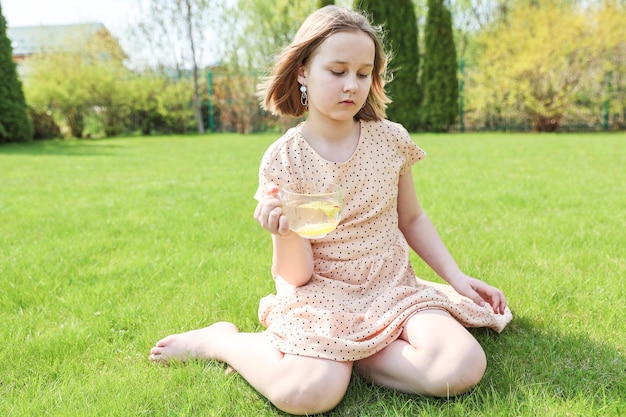 A beautiful happy girl in a beautiful dress is sitting on the grass in the park with a cup of herbal green tea on a hot summer day Healthcare or herbal medicine concept