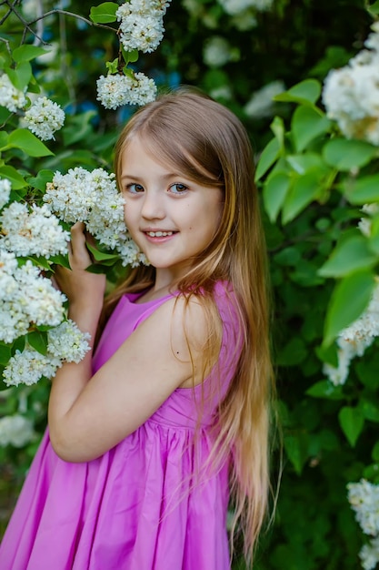 a beautiful happy longhaired blonde girl in a pink dress is standing by a white lilac bush