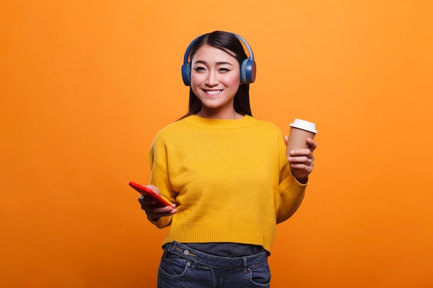 Beautiful happy positive asian woman wearing modern wireless headphones while having a cup of coffee. realxed smiling heartily attractive young adult person enjoying music on smartphone device.