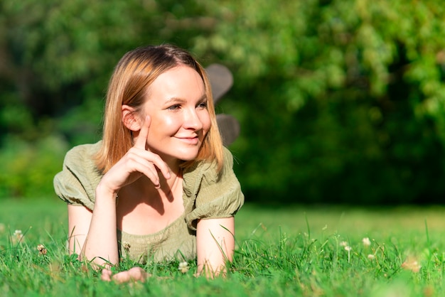 Beautiful happy positive girl, young cheerful woman is lying on her chest on green grass in the park or forest and smiling, enjoying walking, good sunny warm hot weather, looking away, copy space