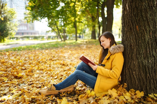 The beautiful happy smiling brown-haired woman in yellow coat and jeans sitting under the maple tree with a red book in fall city park on a warm day. Autumn golden leaves. Reading concept