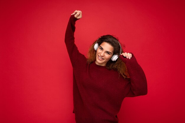 Beautiful happy smiling young brunette curly woman wearing dark red sweater isolated over red background wall wearing white bluetooth headphones listening to music and having fun looking at camera.