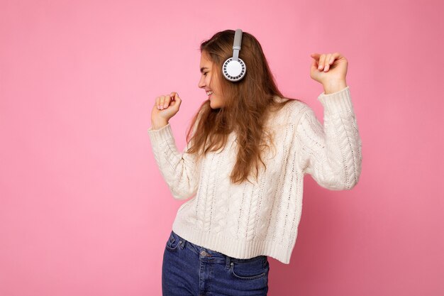 Beautiful happy smiling young brunette curly woman wearing white sweater isolated on pink background wall wearing white bluetooth headphones listening to cool music looking to the side and moving.