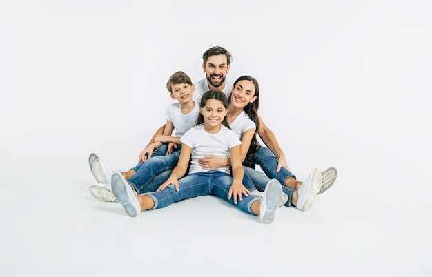 Beautiful and happy smiling young family in white T-shirts are hugging and have a fun time together while sitting on the floor and looking on camera