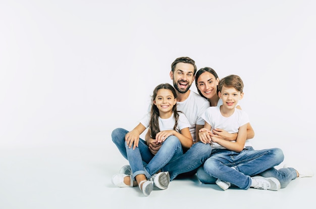 Beautiful and happy smiling young family in white T-shirts are hugging and have a fun time together while sitting on the floor and looking on camera