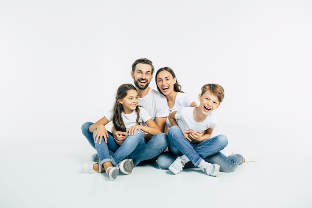 Beautiful and happy smiling young family in white T-shirts are hugging and have a fun time together while sitting on the floor and looking on camera