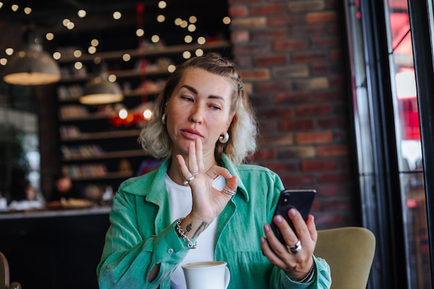 Beautiful happy stylish woman in green shirt sitting in loft cafe drinking cappuccino making video call with her cell phone and showing ok sign