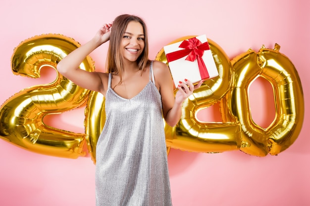 Beautiful happy woman holding red gift box in front of 2020 christmas balloons isolated over pink