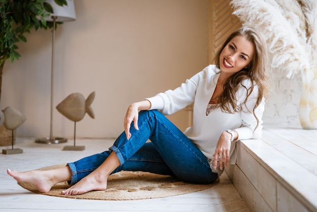 Beautiful happy woman sitting by the window on the floor in jeans and a light blouse