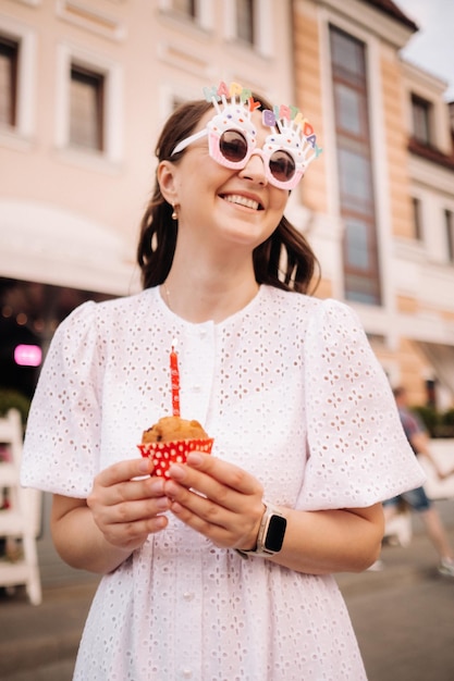 A beautiful happy woman in a white dress holds a cake in her hands on the street of the city celebrating her birthday