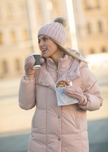 Beautiful happy young adult woman drinking coffee wearing winter clothes and smiling. Beautiful woman holding paper coffee cup outdoors in the city.