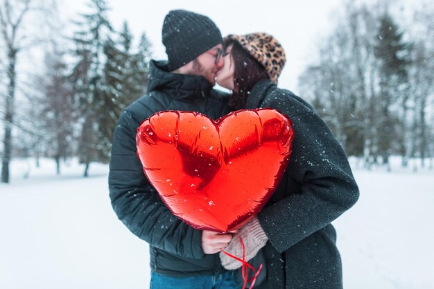 Beautiful happy young couple of lovers holding a red heart balloon and kissing in a winter park with snow Feelings love and relationships