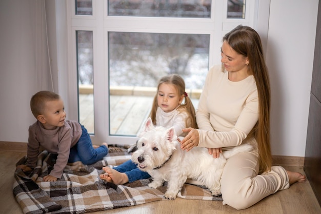 A beautiful and happy young mother and her children son and daughter caress a Dog of the West Highland White Terrier breed at home by the window. Comfort. Warm tones. High quality photo