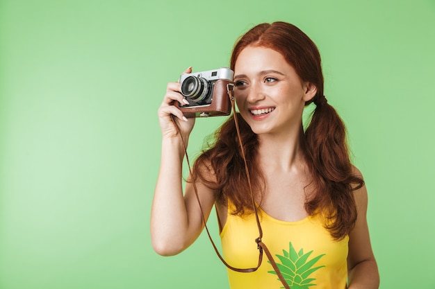 beautiful happy young redhead girl photographer posing isolated over green wall background holding camera.