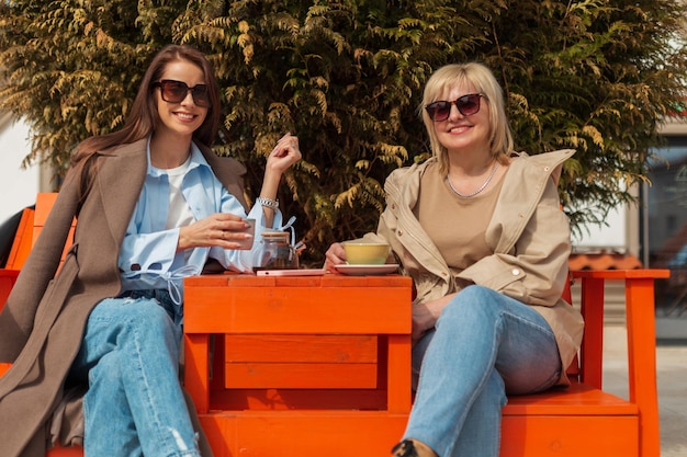 Beautiful happy young woman daughter and happy older woman in trendy fashion clothes with sunglasses are sitting outdoors in a cafe and drinking tea on a sunny spring day