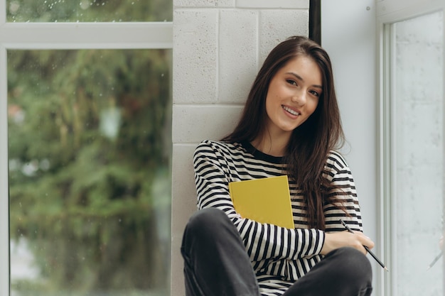 A beautiful happy young woman is sitting on the windowsill with a book in her hands