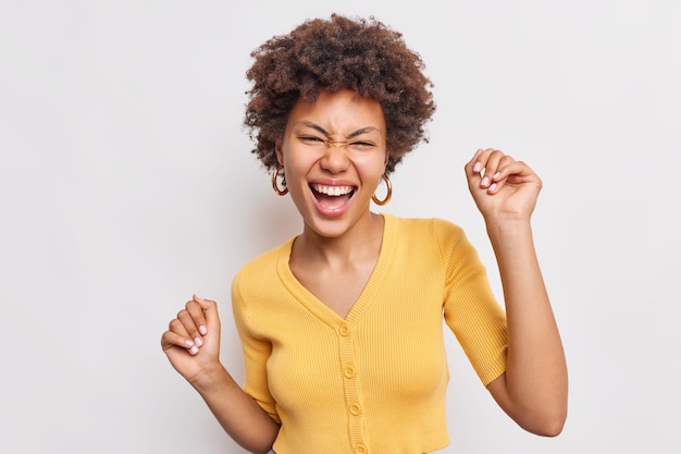 Beautiful happy young woman with curly hiar feels free and pleased raises hands up has fun dances joyful against white wall