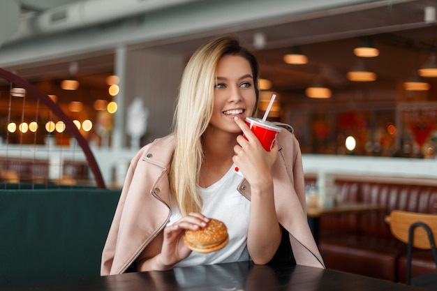 Beautiful happy young woman with a hamburger and drink eating fast food