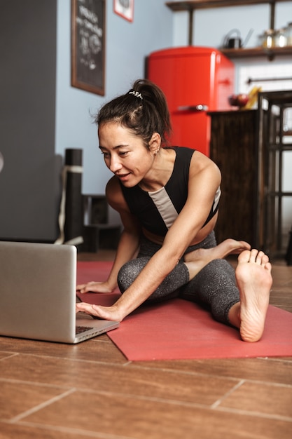 Beautiful healthy woman doing yoga exercises while sitting on a fitness mat at home, using laptop computer, stretching