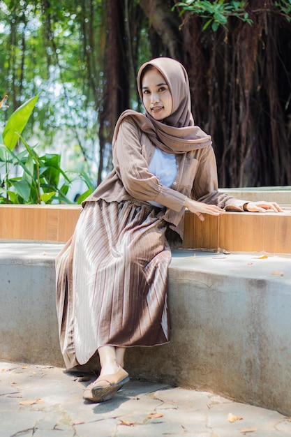 Photo a beautiful hijab indonesia woman smiles at the camera leaning back and sitting on a chair in an out