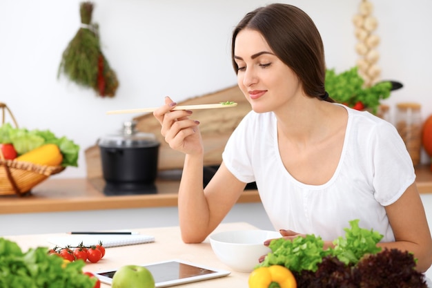 Beautiful  Hispanic woman is cooking in the kitchen. Housewife is tasting fresh salad while sitting at the table