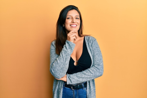 Beautiful hispanic woman wearing casual clothes smiling looking confident at the camera with crossed arms and hand on chin. thinking positive.