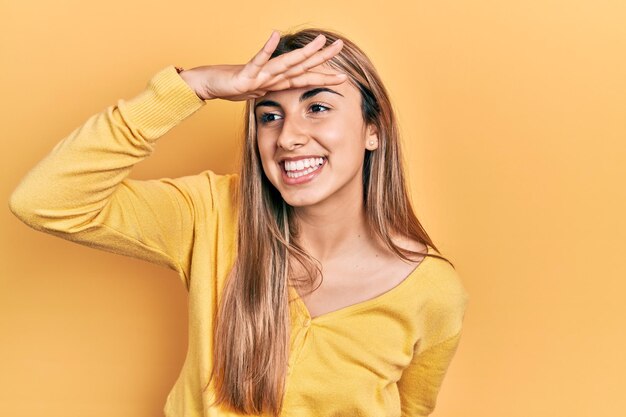 Beautiful hispanic woman wearing casual yellow sweater very happy and smiling looking far away with hand over head searching concept