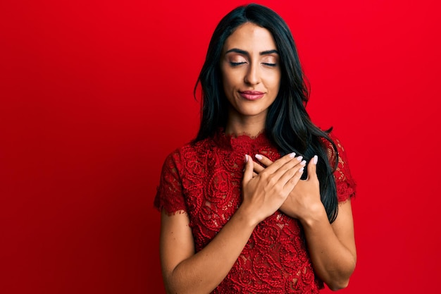 Beautiful hispanic woman wearing elegant clothes over red background smiling with hands on chest with closed eyes and grateful gesture on face health concept