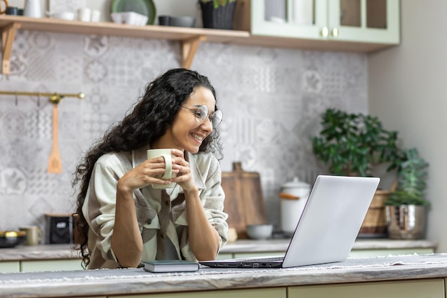 Beautiful hispanic woman with curly hair at home sitting in kitchen and watching video content on