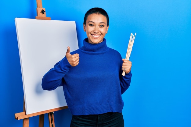 Beautiful hispanic woman with short hair holding brushes close to easel stand smiling happy and positive thumb up doing excellent and approval sign