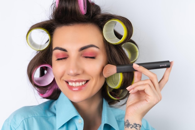 Beautiful housewife Young cheerful woman with hair curlers bright pink makeup and a cotouring brush is preparing for a date night dinner on a white background