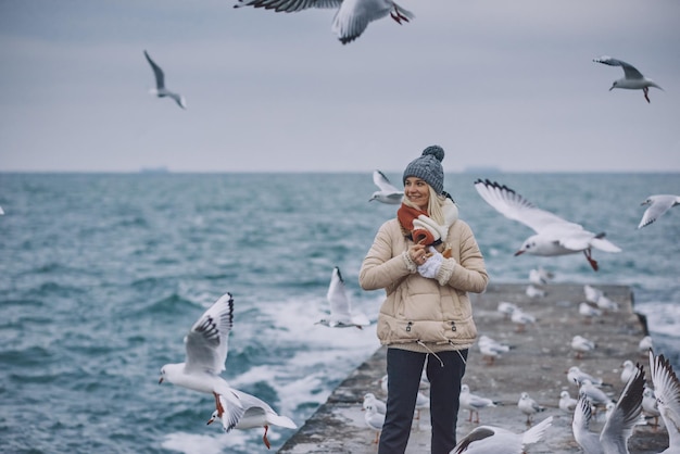 Beautiful image of young smiling woman feeds seagulls on the sea Pretty female wearing coat scarf hat watching flying seagulls by the sea on cold season People and nature concept