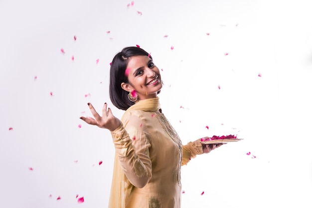 Beautiful Indian or Asian young girl in traditional wear throwing rose petals from plate, standing isolated over white background