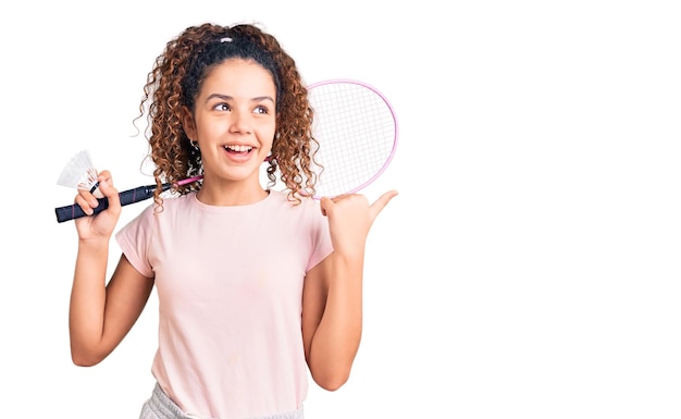 Beautiful kid girl with curly hair holding badminton racket and shuttlecock pointing thumb up to the side smiling happy with open mouth