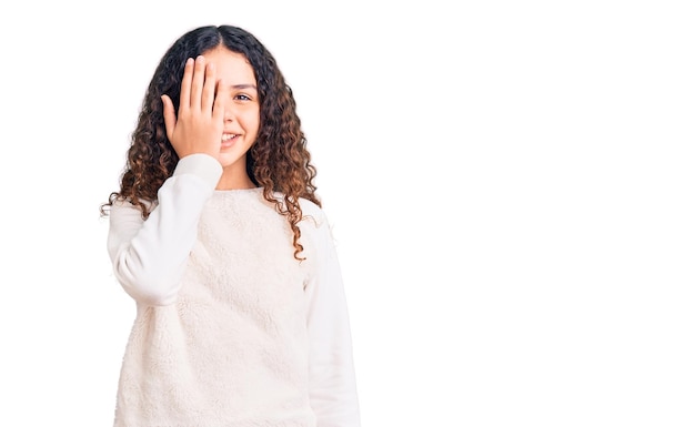 Beautiful kid girl with curly hair wearing casual clothes covering one eye with hand, confident smile on face and surprise emotion.