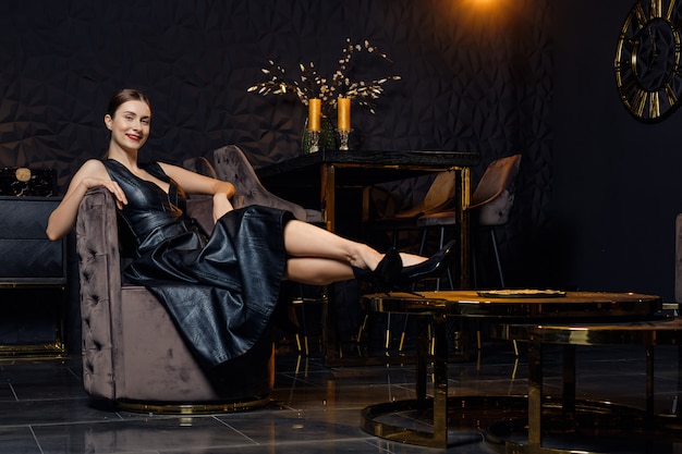 Beautiful lady in leather dress relax in cosy armchair in dark room