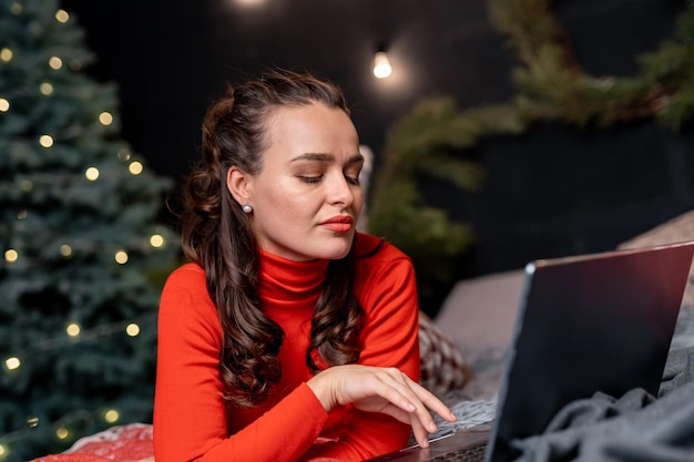 Beautiful lady in red clothes lying in front of her laptop Woman with notebook on the plaid near Christmas tree Woman with curly hair working on her laptop near fir tree