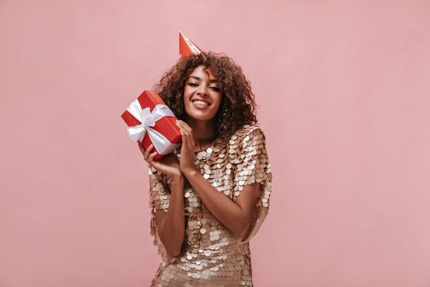 Beautiful lady with curly hair in beige dress and holiday cap smiling and holding red gift box on isolated pink wall..