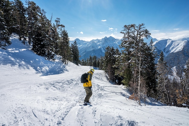 Beautiful landscape of the Arkhyz ski resort with mountains snow forest and man snowboarder in yellow jacket and backpack on a sunny winter day Caucasus Mountains Russia