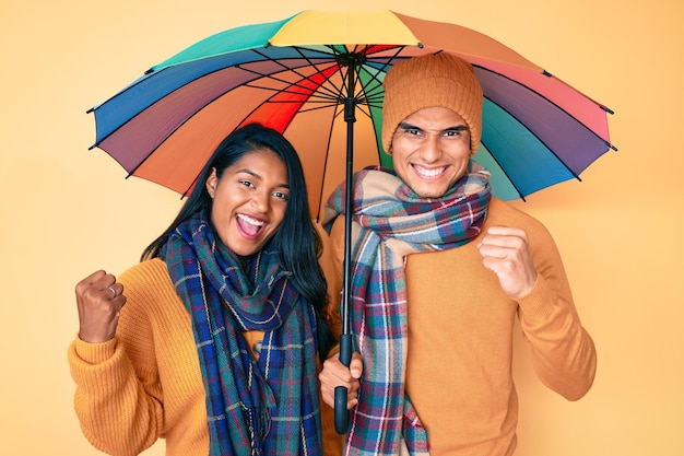 Beautiful latin young couple under colorful umbrella screaming proud, celebrating victory and success very excited with raised arms