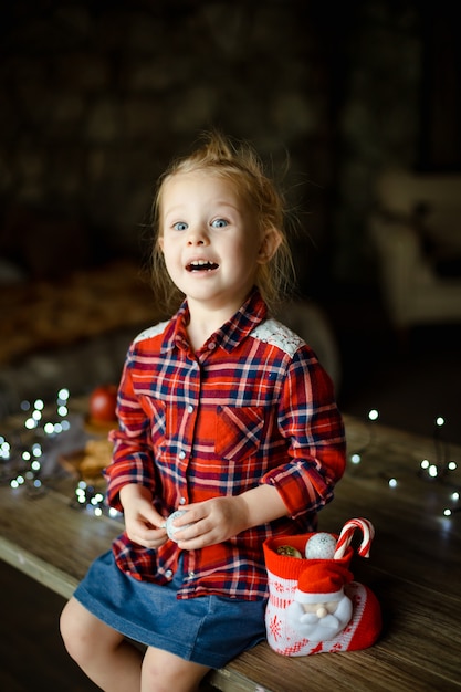 A beautiful little blonde in a traditional plaid shirt opens a chocolate bar from her sweet Christmas gift