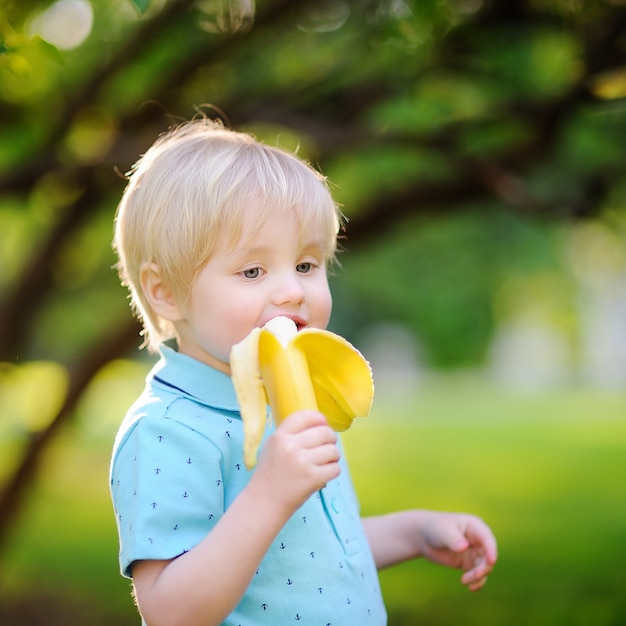 Beautiful little boy eating banana during picnic in summer sunny park