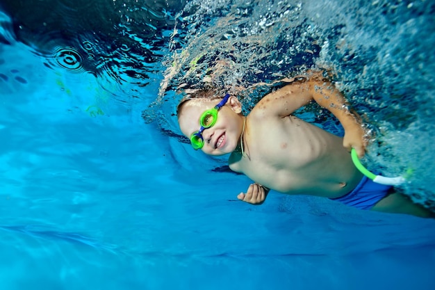 A beautiful little boy is swimming under the water in the pool looking forward and smiling