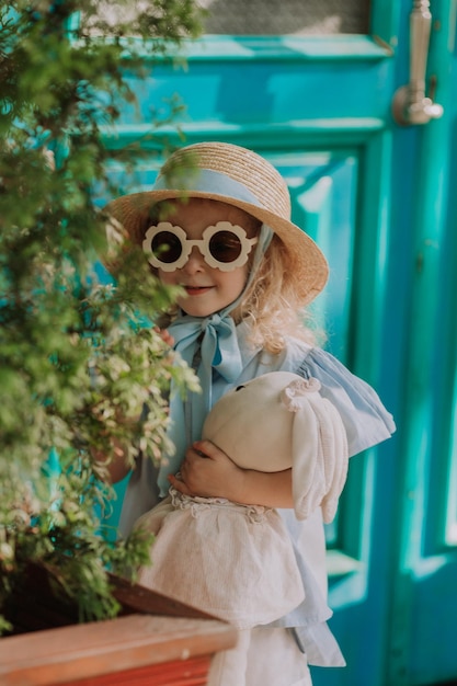 beautiful little girl in blue dress, straw hat and sunglasses playing outdoor, blue background, card