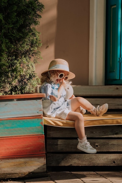 beautiful little girl in blue dress, straw hat and sunglasses playing outdoor, blue background, card