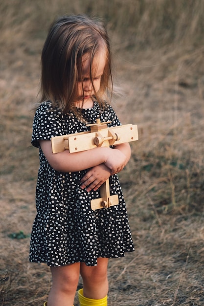 A beautiful little girl in a dress holds a wooden airplane in her hands against the background of autumn nature and sunset Retro style Ideas for postcards posters Children's dreams and fantasies