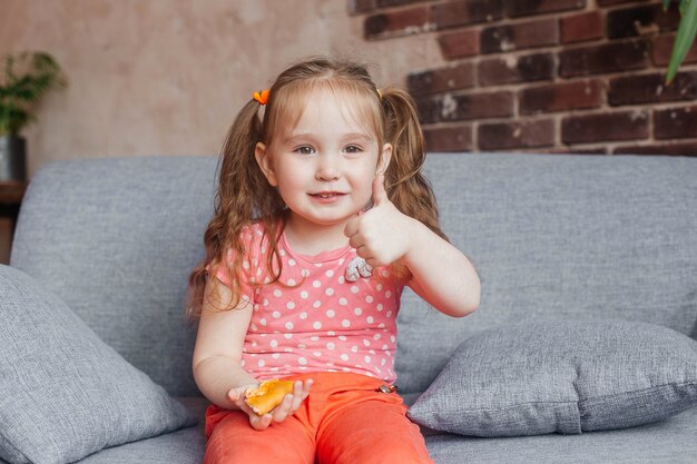 Beautiful little girl eating pizza at home and raising thumb up